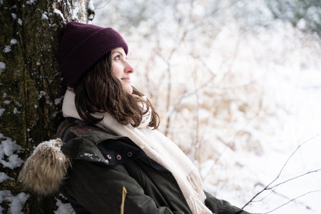 A young girl walks in a beautiful winter forest among tall trees, firs, covered with snow. Magnificent nature and views. Fashionable image, clothes, parka, hat, mittens, blue jeans.の写真素材