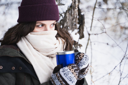 Young girl, woman in knitted mittens drinks tea, drinks coffee from thermo mug. Walking in beautiful...