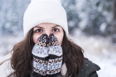 Young girl, woman in knitted mittens feels cold. Walking in beautiful winter forest among trees, firs, covered with snow. Magnificent nature. Fashionable image, clothes, parka, hat, blue jeans.の写真素材