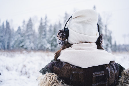 Young girl, woman in knitted mittens listening to music on headphones. Walking in beautiful winter forest among trees, firs, covered with snow. Magnificent nature. Fashionable image, clothes, parka, hat.の写真素材
