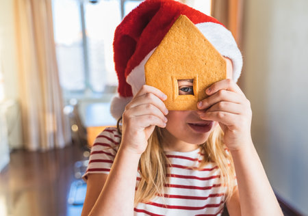 Little girls, sisters in red santa claus hats make christmas gingerbread house. Covered by white glaze, cream with walls, roof, windows, chimney. Decoration with bright multicolored sweets, lollipops.の写真素材