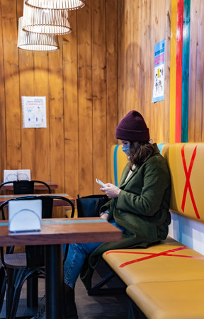 Young girl, woman in protective face mask visits bright cafe. Safe social distance. Stylish image,coat,hat, glasses.Marked prohibited areas,seats for restaurant visitors.Pandemicの写真素材