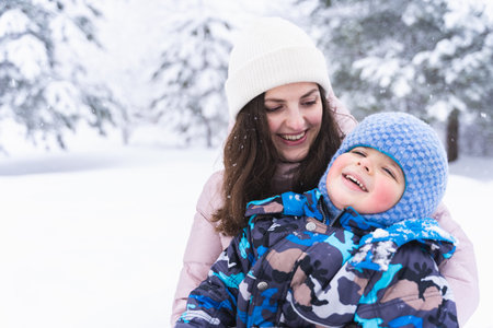 Mother hugs little smiling, laughing cute boy,kid.Red blush on cheeks.Happy family walking in beautiful winter forest,park covered with snow.Blue hat helmet,bright warm khaki jacket.Fashionable image.の写真素材