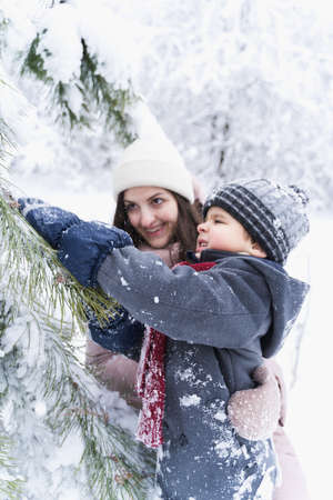 Mom kissing little stylish smiling happy cute boy, kid. Lovely family walking,activity in beautiful winter forest, park.Trees covered with snow.Gray hat,woolen coat,jacket,red scarf.Fashionable image.の写真素材