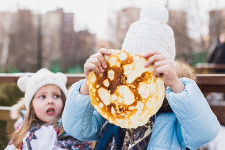 Russian national festival Maslenitsa, shrovetide. Little cute beautiful European girls in headscarf eat big tasty pancakes and have fun on winter pancake week. Traditional folf slavic street carnival.の写真素材