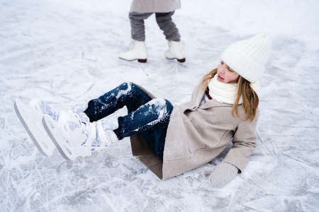 Beautiful little girls, sisters learn to skate on ice skating rink in park. Fall and have fun. Stylish looks, warm woolen coats, white hats, scarves, snoods. Winter family activities, games outdoors.の写真素材