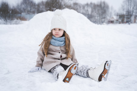Beautiful little cute girl learn to skate on ice skating rink in park. Fall down and have fun. Stylish look, warm woolen coat, white hat, scarf, snood. Winter family activities, sport, games outdoors.の写真素材