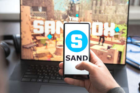 Russia Moscow 30.05.2021.Logo, screenshot of blockchain nft ethereum cryptocurrency game Sandbox in laptop, mobile phone.Man playing with crypto coins, token sand.Earning digital money.Lands, heroes.のeditorial素材