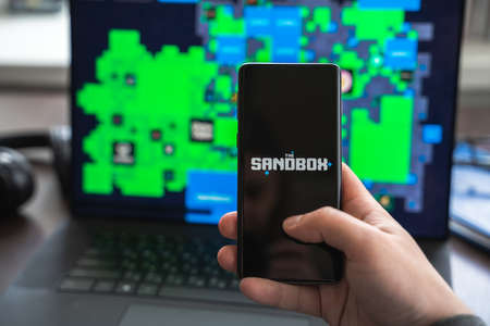 Russia Moscow 30.05.2021.Logo,screenshot of blockchain nft ethereum cryptocurrency game Sandbox in laptop,mobile phone.Man playing with crypto coins,token sand.Earning digital money.Lands,heroes.のeditorial素材