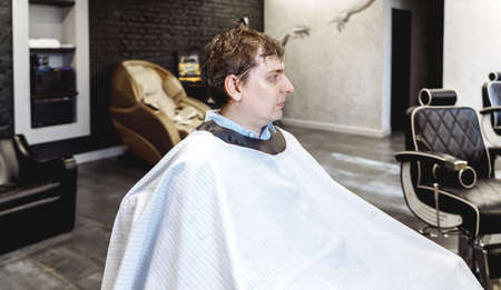 Visit to the barbershop.Young stylish man,businessman makes fashionable haircut.Barber, hairdresser,stylist and client,customer sitting in dark men's beauty salon with black chair covered with a cape.の写真素材