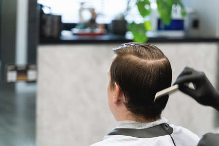 Visit to the barbershop. Young stylish man makes fashionable haircut. Barber,hairdresser,stylist combing hair and client,customer sitting in dark men's beauty salon with black chair covered with cape.の写真素材