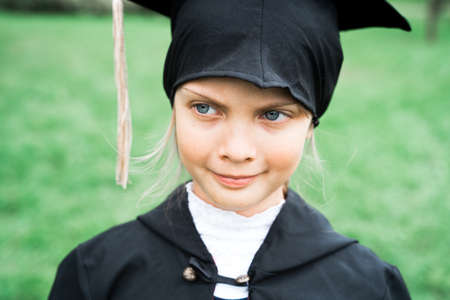 Back to elementary school concept. Little girl goes to first grade. Ceremony of graduating. Black gown, academic cap with tassels. Party with parents, teachers in park, yard. Last day, end of year.の写真素材