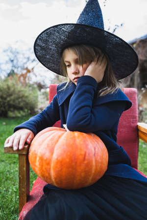 Scary little girl in witch costume, hat with big pumpkin celebrating halloween holiday. Sitting on armchair in coat with pumpkin. Stylish image. Horror, fun at children's party in barn on street.の写真素材