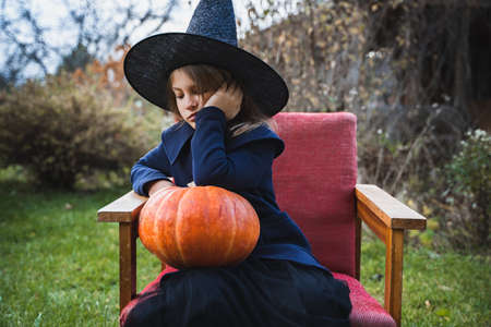 Scary little girl in witch costume, hat with big pumpkin celebrating halloween holiday. Sitting on armchair in coat with pumpkin. Stylish image. Horror, fun at children's party in barn on street.の写真素材