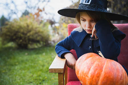 Scary little girl in witch costume, hat with big pumpkin celebrating halloween holiday. Sitting on armchair in coat with pumpkin. Stylish image. Horror, fun at children's party in barn on street.の写真素材