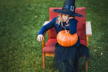 Scary little girl in witch costume, hat with big pumpkin celebrating halloween holiday. Sitting on armchair in coat with pumpkin. Stylish image. Horror, fun at children's party in barn on street.の写真素材