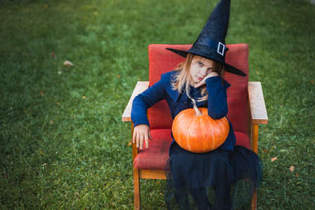 Scary little girl in witch costume, hat with big pumpkin celebrating halloween holiday. Sitting on armchair in coat with pumpkin. Stylish image. Horror, fun at children's party in barn on street.の写真素材
