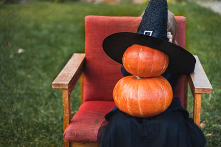 Scary little girl in witch costume, hat with many pumpkins celebrating halloween holiday. Sitting on armchair in coat with pumpkin. Stylish image. Horror, fun at children's party in barn on street.の写真素材