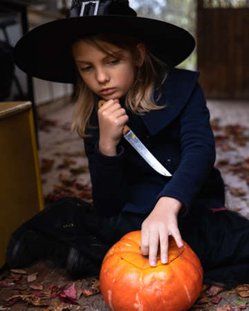 Little girls make jack-o-lantern from big pumpkins for celebratiion of halloween holiday.Witch costume, hat, coat. Cut with knife,take out pulp with seeds.Outdoors activity, backyard.Children's party.の写真素材