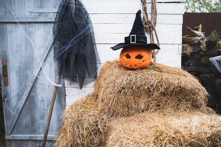 Orange pumpkin for halloween in black witch, wizard hat, jack-o-lantern with scary carved eyes, mouth. Hay, haystack near barn. DIY home, street decoration, entertainment for children, horror.の写真素材