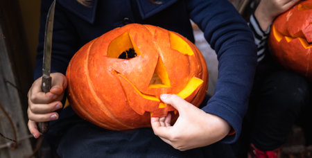Little girls make jack-o-lantern from big pumpkins for celebratiion of halloween holiday.Witch costume, hat, coat. Cut with knife,take out pulp with seeds.Outdoors activity, backyard.Children's party.の写真素材