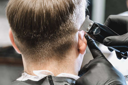 Visit to the barbershop. Hairdresser, barber shaves client's temples,sides with hair clipper. Young stylish man makes fashionable haircut.Customer sitting in dark men's beauty salon covered with cape.の写真素材