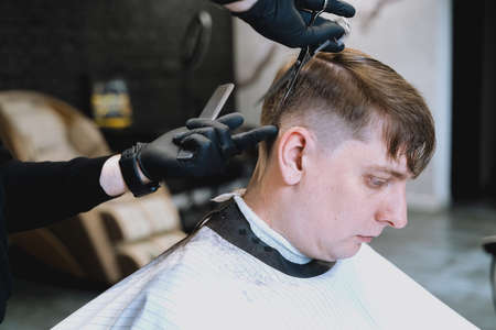 Visit to barbershop. Stylish man makes fashionable haircut. Barber, hairdresser, stylist with scissors, comb cutting,combing hair. Client,customer sitting in dark men's beauty salon covered with cape.の写真素材