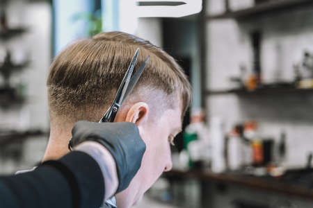 Visit to barbershop. Stylish man makes fashionable haircut. Barber, hairdresser, stylist with scissors, comb cutting, combing hair. Client, customer sitting in dark men's beauty salon covered with cape.の写真素材