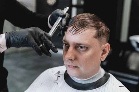 Visit to barbershop. Stylish man makes fashionable haircut. Barber, hairdresser, stylist with scissors, comb cutting, combing hair. Client, customer sitting in dark men's beauty salon covered with cape.の写真素材