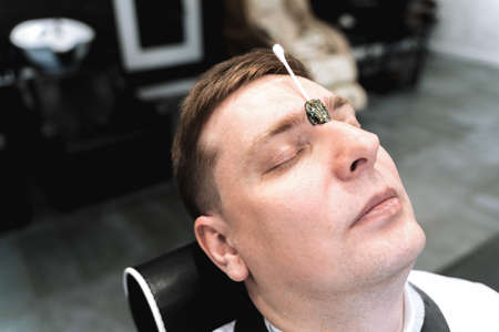 Visit to barbershop.Wax depilation,hair removal between the eyebrows.Stylish man makes fashionable haircut.Barber,hairdresser,stylist.Client,customer sits in dark men's beauty salon covered with cape.の写真素材