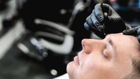 Visit to barbershop.Plucking eyebrows with tweezers,hair removal.Stylish man makes fashionable haircut.Barber,hairdresser,stylist.Client,customer sits in dark men's beauty salon covered with cape.の写真素材