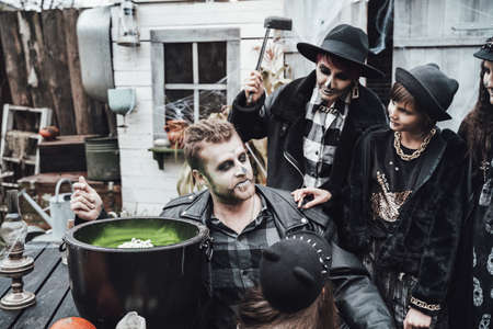 Scary family, mother, father, daughters celebrating halloween. Cauldron of potion.Terrifying black skull half-face makeup,witch costumes,stylish images.Horror,fun at children's party in barn on street.の写真素材