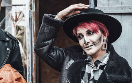Scary woman, girl celebrating halloween.Terrifying black skull half-face skeleton makeup and witch costume in stylish image. Jacket, hat. Horror, fun at photoshoot, holiday party near barn on street.の写真素材