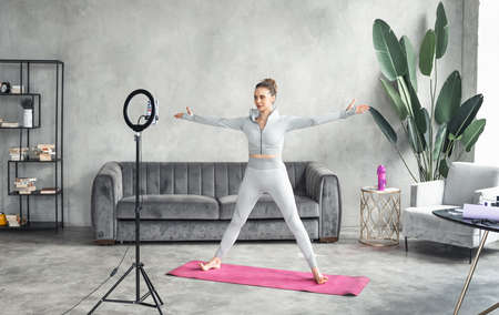Sporty slim woman yoga coach, fitness instructor records video of online yoga training in mobile phone camera. Professional blogger vlogger. Workout for social net. Exercises at home on fitness mat.の写真素材