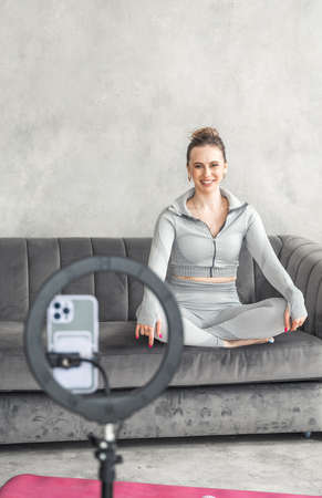 Sporty slim woman yoga coach, fitness instructor records video of online yoga training in mobile phone camera. Professional blogger vlogger. Workout for social net. Exercises at home on fitness mat.の写真素材