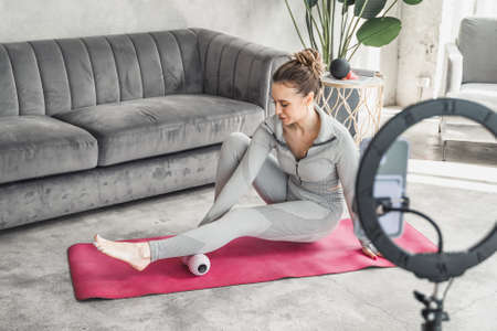 Sporty slim woman yoga coach, fitness instructor records video of online yoga training in mobile phone camera. Professional blogger vlogger. Workout for social net. Exercises at home on fitness mat.の写真素材