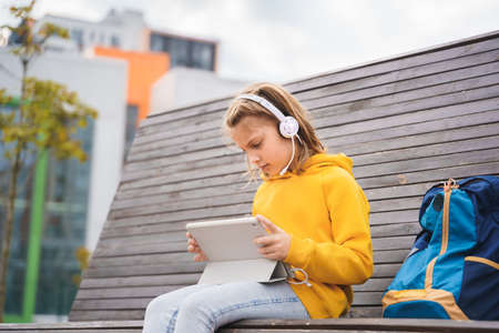 Girl in headphones, headset with tablet. Study, online conference, lesson, education. Remote learning on street bench in school yard, park. School girl with backpack. Chat, talk with friends, teacher.の写真素材