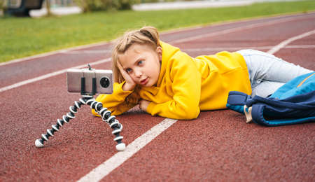 Back to elementary, primary school. Little girls with backpack,mobile phone.Blog, blogger vlogger recording video on sport playgroumd stadium.Chat talk with mom friends classmates on break. Education for kids. using technologies.の写真素材