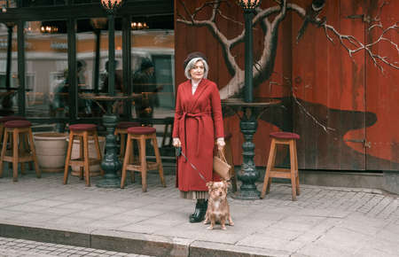 Elderly senior woman walking with lovely dog pet in red coat. Old retired aged lady on walk on city street with flowers.Stylish elder person pensioner. Quite calm happy years in metropolis.の写真素材