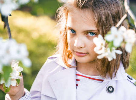 Little girl is walking in spring garden with apple trees and white flowers. Romantic stylish spring kid in coat. Blooming in sunny park. beautiful nature photography. Spring bloom enjoy, relax.の写真素材