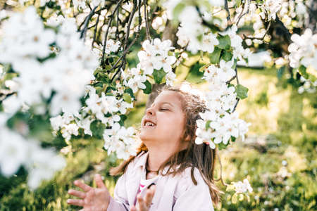 Little girl is walking in spring garden with apple trees and white flowers. Romantic stylish spring kid in coat. Blooming in sunny park. beautiful nature photography. Spring bloom enjoy, relax.の写真素材