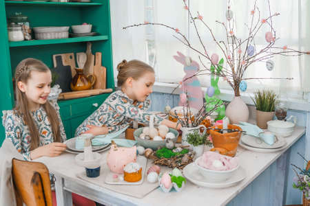 Girls, sisters at table with Easter decoration. Celebration in kitchen.Tablescape for home Easter holiday.Family religious traditional festive food, meal. Colored eggs, cake, funny bunny, candy sweet.の写真素材
