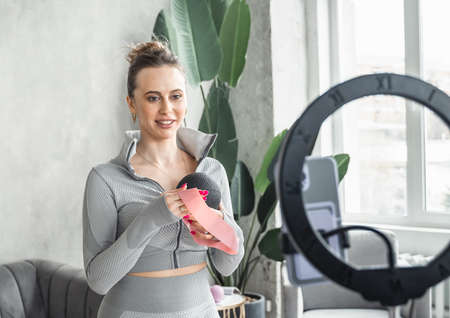 Sporty slim woman yoga coach, fitness instructor records video of online yoga training in mobile phone camera. Professional blogger vlogger. Workout for social net. Exercises at home on fitness mat.の写真素材