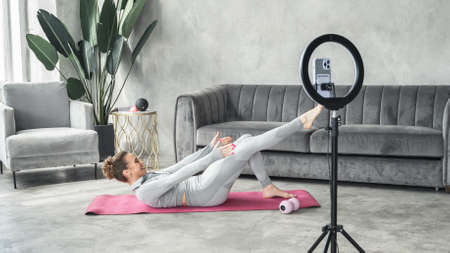 Sporty slim woman yoga coach, fitness instructor records video of online yoga training in mobile phone camera. Professional blogger vlogger. Workout for social net. Exercises at home on fitness mat.の写真素材