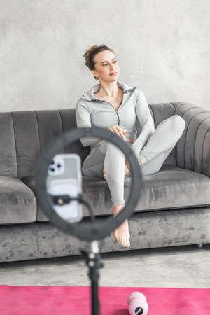 Sporty slim woman yoga coach, fitness instructor records video of online yoga training in mobile phone camera. Professional blogger vlogger. Workout for social net. Exercises at home on fitness mat.の写真素材