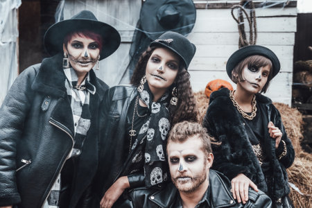 Scary family, mother, father, daughters celebrating halloween. Terrifying black skull half-face makeup and witch costumes, stylish images.Horror,fun at children's party in barn on street.Hats,jackets.の写真素材