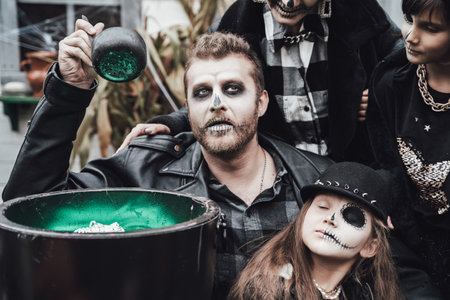 Scary family, mother, father, daughters celebrating halloween. Cauldron of potion.Terrifying black skull half-face makeup,witch costumes,stylish images.Horror,fun at children's party in barn on street.の写真素材
