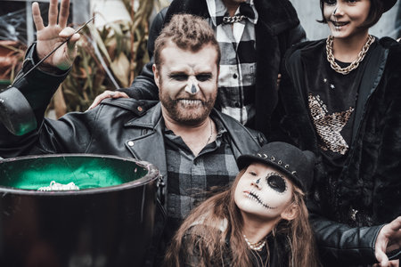 Scary family, mother, father, daughters celebrating halloween. Cauldron of potion.Terrifying black skull half-face makeup,witch costumes,stylish images.Horror,fun at children's party in barn on street.の写真素材