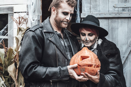 Scary love family couple man,woman celebrating halloween with pumpkin jack-o-lantern.Terrifying black skull half-face makeup,witch costumes,stylish images,jacket,hat. Fun,holiday party at street barn.の写真素材