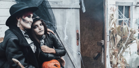 Scary family,mother,daughter celebrating halloween. street barn. Pumpkin jack-o-lantern. Terrifying black skull half-face makeup and witch costumes, broom, stylish images hat, jackets. children's party.の写真素材
